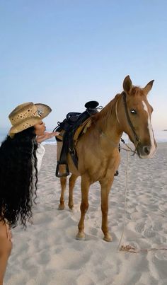 a woman in a cowboy hat is standing next to a horse on the beach,