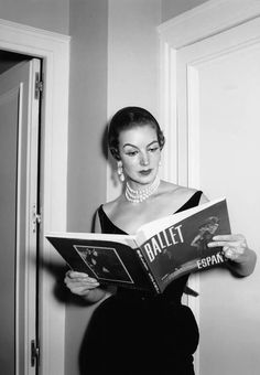 a black and white photo of a woman reading a ballet program book in front of a door