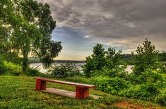 a wooden bench sitting on top of a lush green field next to a river under a cloudy sky