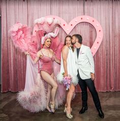 a man and woman posing for a photo in front of a heart shaped backdrop with pink feathers