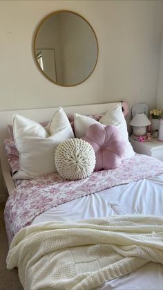 a white bed topped with lots of pillows next to a wall mounted mirror and dresser