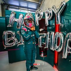 a little boy in a race suit standing next to some balloons that say happy birthday