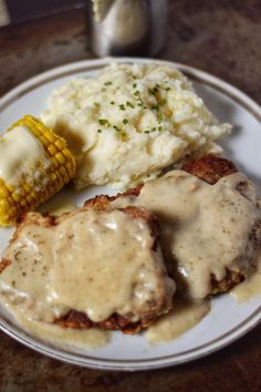 a white plate topped with meat covered in gravy and corn on the cob