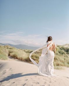 a woman in a white dress is walking on the sand with a veil over her head