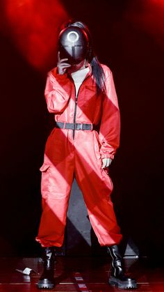 a woman in a red jumpsuit on stage with her hands to her face and one hand behind her head