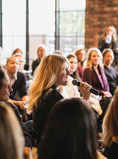 a woman speaking into a microphone in front of a group of people sitting on the floor