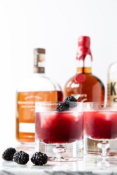 two glasses filled with red liquid and blackberries next to bottles of alcohol on a table