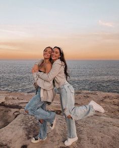 two women hugging each other while standing on rocks near the ocean at sunset or sunrise