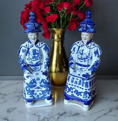 two blue and white figurines sitting next to a vase with red flowers in it