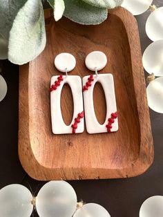 two white and red earrings sitting on top of a wooden tray next to a plant