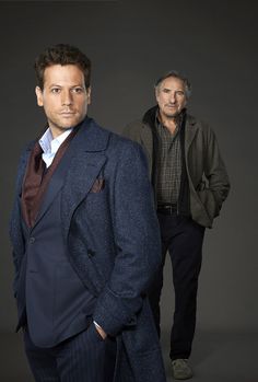 two men standing next to each other in front of a dark background with one man wearing a suit and the other looking at the camera