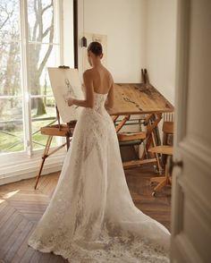 a woman in a white wedding dress is holding an easel and looking out the window