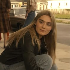 a woman sitting on the ground next to a car and another person standing behind her