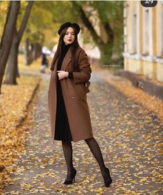 a woman in a brown coat and black hat standing on a leaf covered street with trees