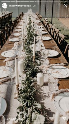 a long wooden table with white plates and greenery on it, surrounded by candles