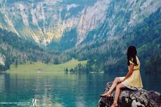 a woman sitting on top of a rock next to a lake with mountains in the background