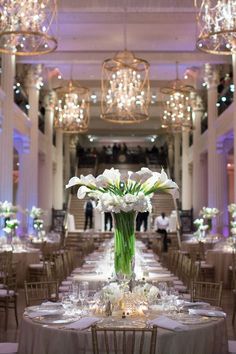 a long table with white flowers in a tall vase on it and chandeliers hanging from the ceiling