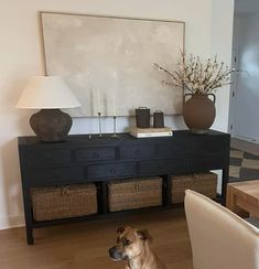 a dog sitting on the floor in front of a table with two baskets and a lamp