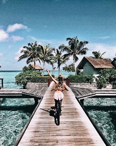 a woman riding a bike down a wooden walkway next to the ocean with palm trees