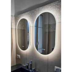 two round mirrors in the middle of a bathroom wall with lights on each side and an illuminated mirror above them