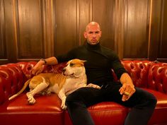 a man sitting on top of a red couch next to a brown and white dog