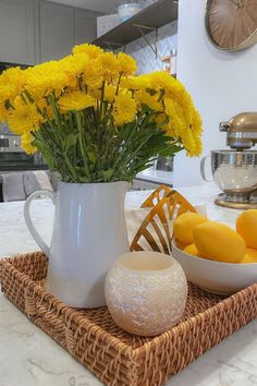 yellow flowers and lemons in a white pitcher on a wicker tray next to a bowl of oranges