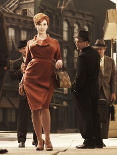 a woman in a brown dress is standing on the street with two men and a suitcase