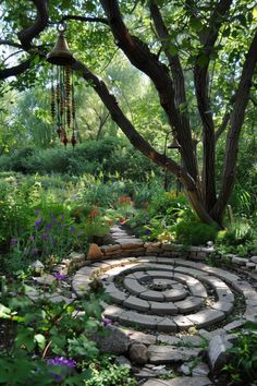 a stone path in the middle of a garden surrounded by trees and flowers, with a wind chime hanging from the tree