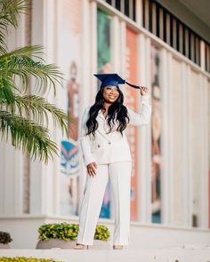 a woman in a white suit and graduation cap poses for a photo outside the building