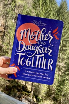 a person holding up a book that says mother and daughter together in front of some trees