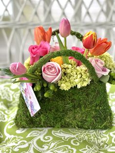 an arrangement of flowers and greenery in a moss covered basket on a tablecloth