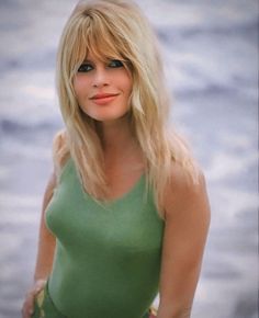 a woman with long blonde hair wearing a green top and posing for a photo in front of the ocean