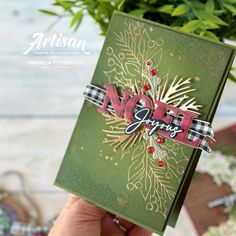 a hand holding up a christmas card with the word noel on it and pine cones