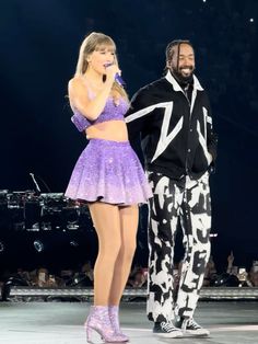 two people standing on stage with one holding a microphone and the other wearing a purple outfit