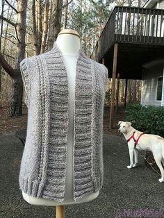 a white dog standing next to a mannequin wearing a gray knitted vest