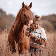 a woman standing next to a brown horse in a field with tall grass and trees