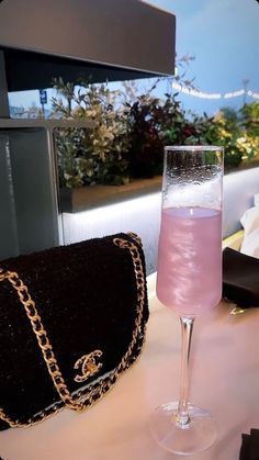 a pink liquid in a wine glass next to a black purse on a white table