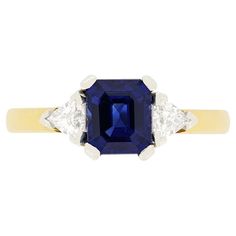 This Vintage trilogy ring from the 1970s centres around a striking 1.20 carat, emerald cut sapphire. Its deep blue hue is flanked by two glittering trillion cut diamonds. Weighing 0.15 carat a piece, each stone is of high quality, with F colour and VS1 clarity. The stones are securely claw set into platinum, enhancing their brilliance. The band is crafted 18 carat yellow gold contrasting beautifully against the cool tones of the sapphire and diamonds. Gemstone: Sapphire Stone Shape: Emerald Cut Carat Weight: 1.20 Metal: 18ct Yellow Gold, Platinum Age: 1970s Size: K 1/2 Diamond Trilogy Ring, Trilogy Ring, Sapphire Stone, Three Stone Rings, The 1970s, Cool Tones, Sapphire Diamond, Three Stone, Emerald Cut