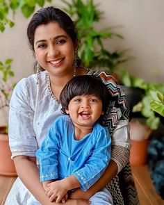 a woman holding a child in her arms and smiling at the camera with potted plants behind her