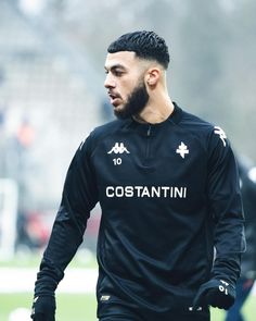 a man in black jersey walking on field with other people behind him wearing gloves and holding soccer balls