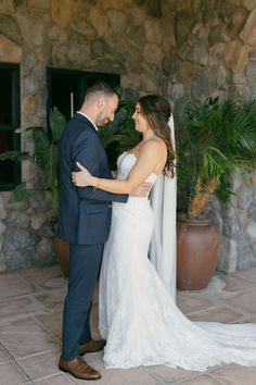 a bride and groom standing next to each other in front of a stone wall with potted plants