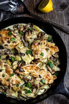 a skillet filled with chicken, mushrooms and artichokes on top of a wooden table