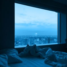 a person is laying in bed looking out the window at the city below them,