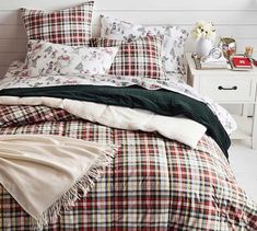 a bed with plaid comforter and pillows on it next to a night stand filled with flowers