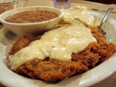 a white plate topped with meat covered in gravy next to a bowl of mashed potatoes