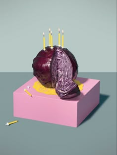 a purple cake with candles on it sitting on top of a pink box in front of a gray background