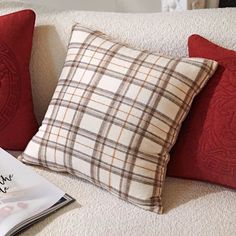 two red pillows sitting on top of a white couch next to an open book and magazine