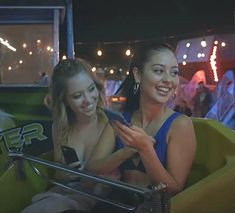 two young women sitting in the back of a green car at night, texting on their cell phones