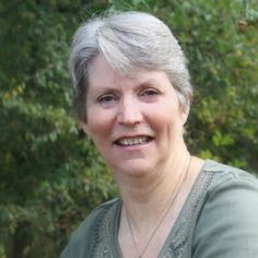 an older woman with gray hair smiling at the camera in front of trees and bushes