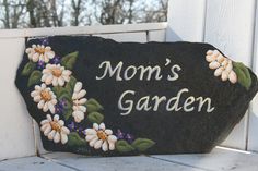 a rock with flowers painted on it that says mom's garden next to a white picket fence